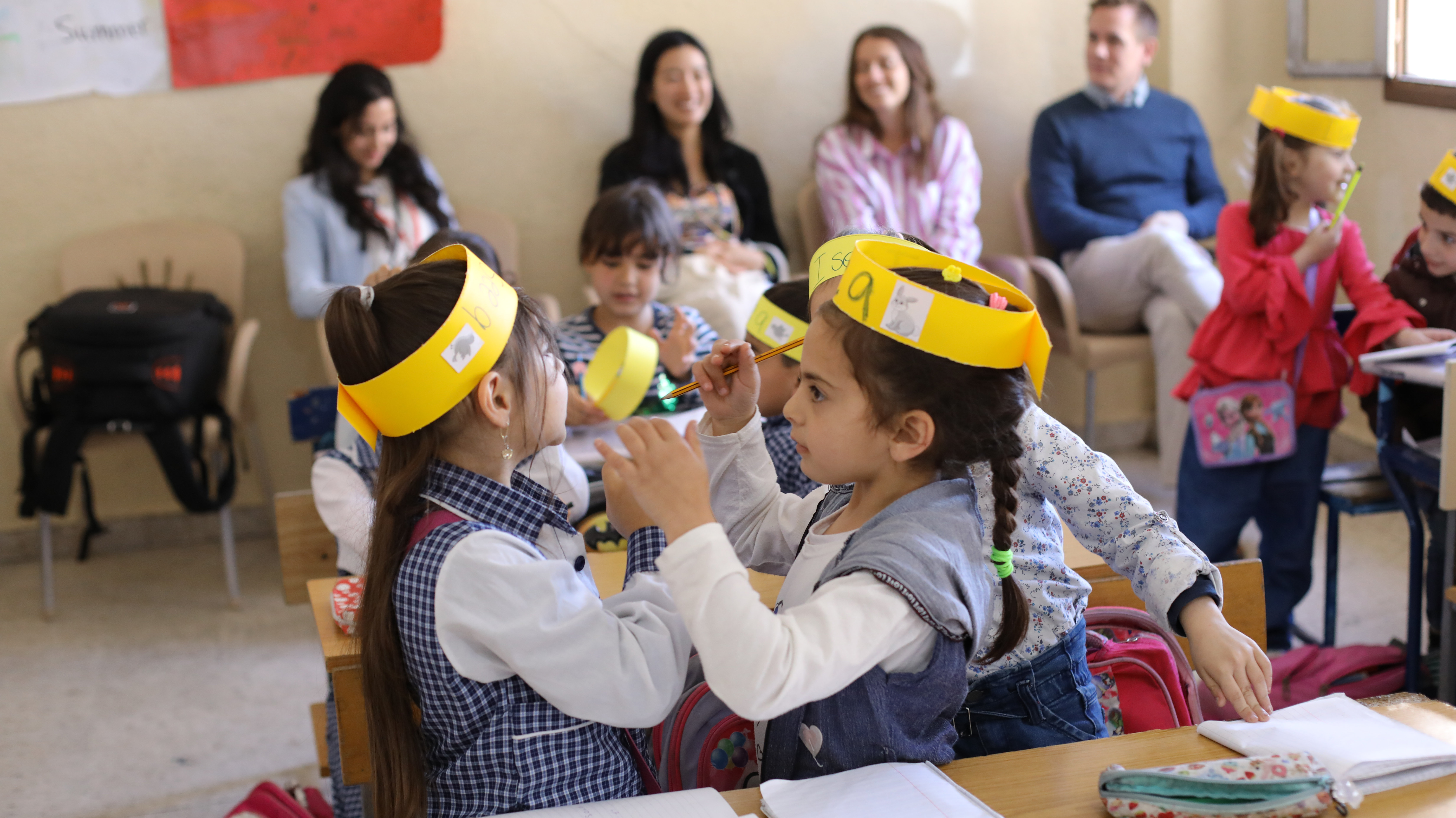 Preview image for the video "Education in Emergencies: Connecting across borders". Preview image for the video "Education in Emergencies: Connecting across borders" - Two young girls in a classroom wearing yellow paper headbands face each other.
