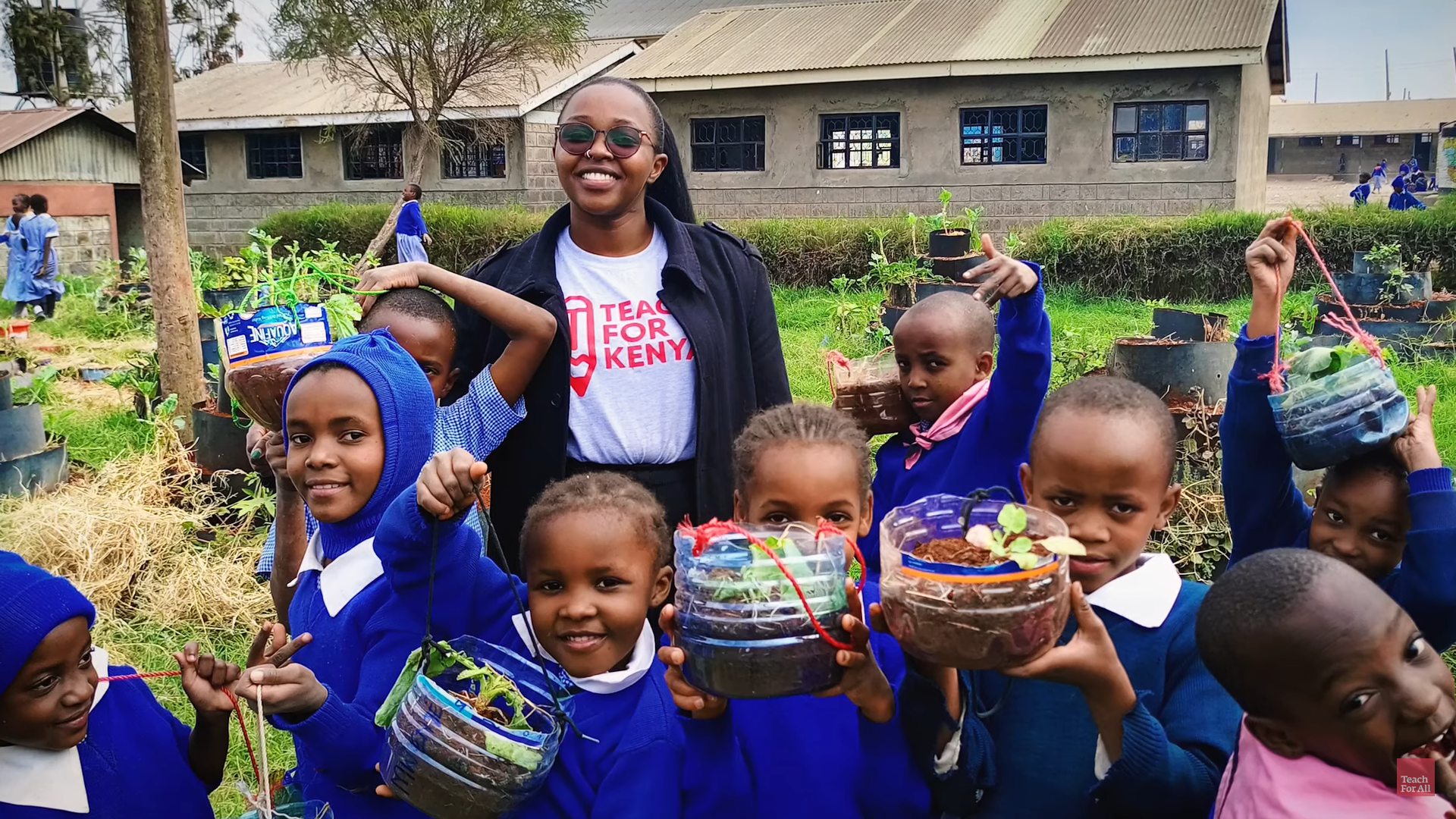 Preview image for the video "Esther Gacigi - Network Changemakers" - A Black female teachers stands in a school yard surrounded by young students holding up plants growing in recycled plastic containers.