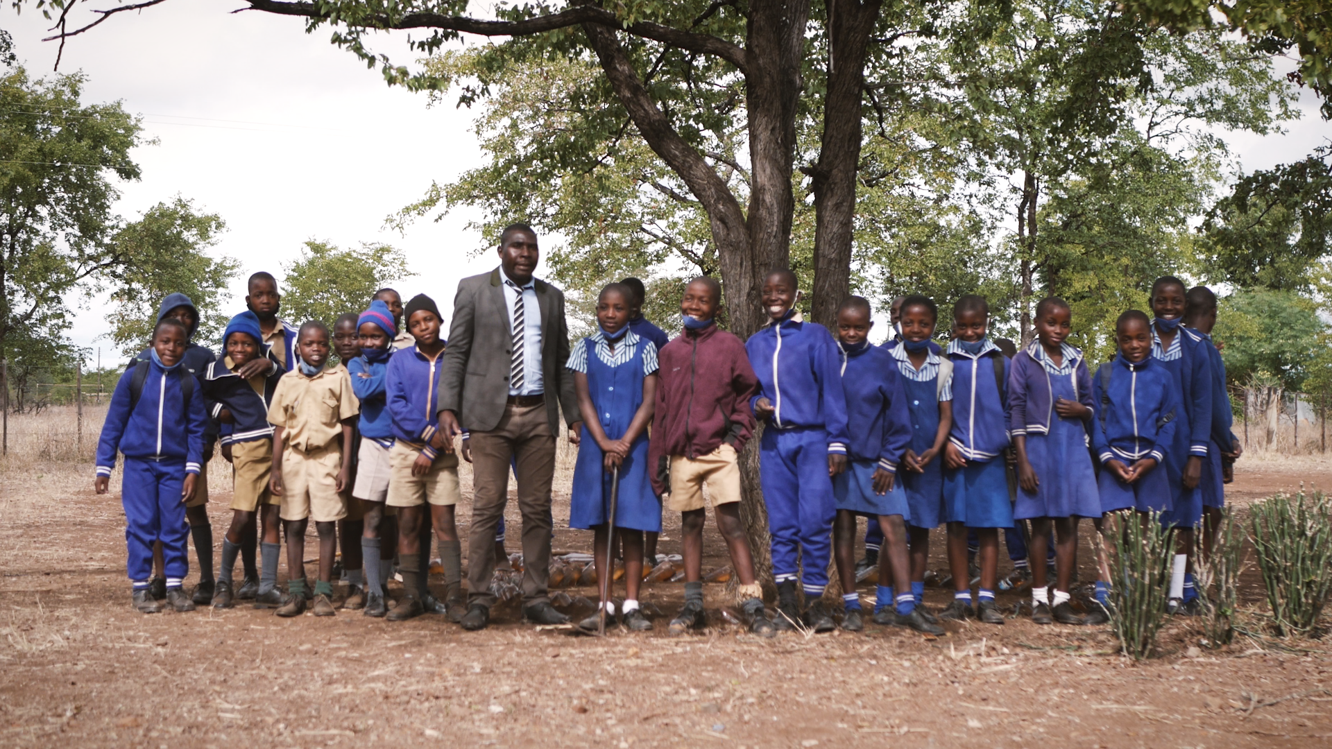 Preview image for the video "Edson Dongo - Teach For Zimbabwe" - In front of a large tree, a tall African man stands in the middle of a row of African students in school uniforms.