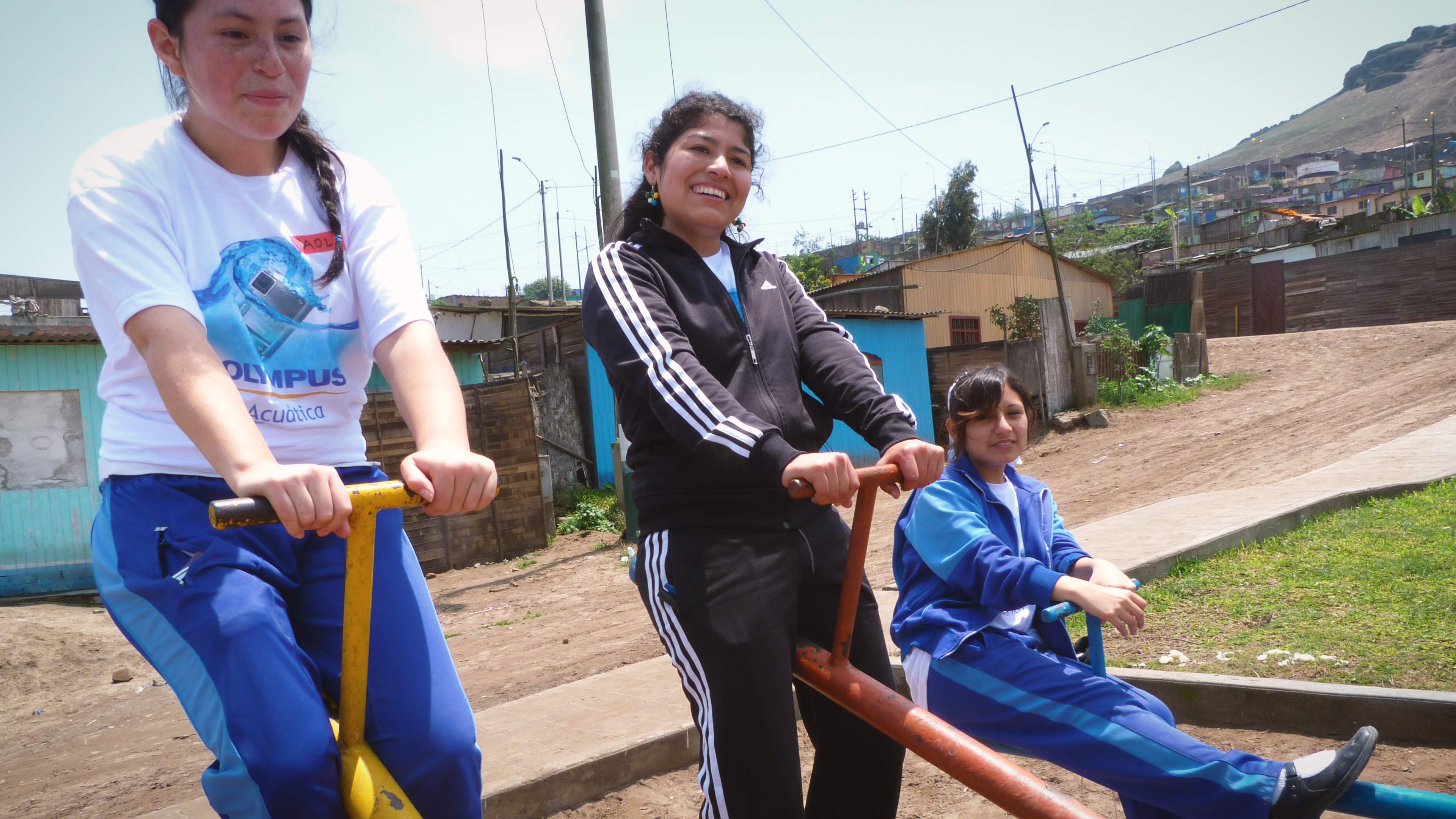 Preview image for the video "Karina Clemente - Enseña Perú" - A young woman in a black track suit smiles as she sits on top of a see saw with smiling girls in blue track suits on either side.