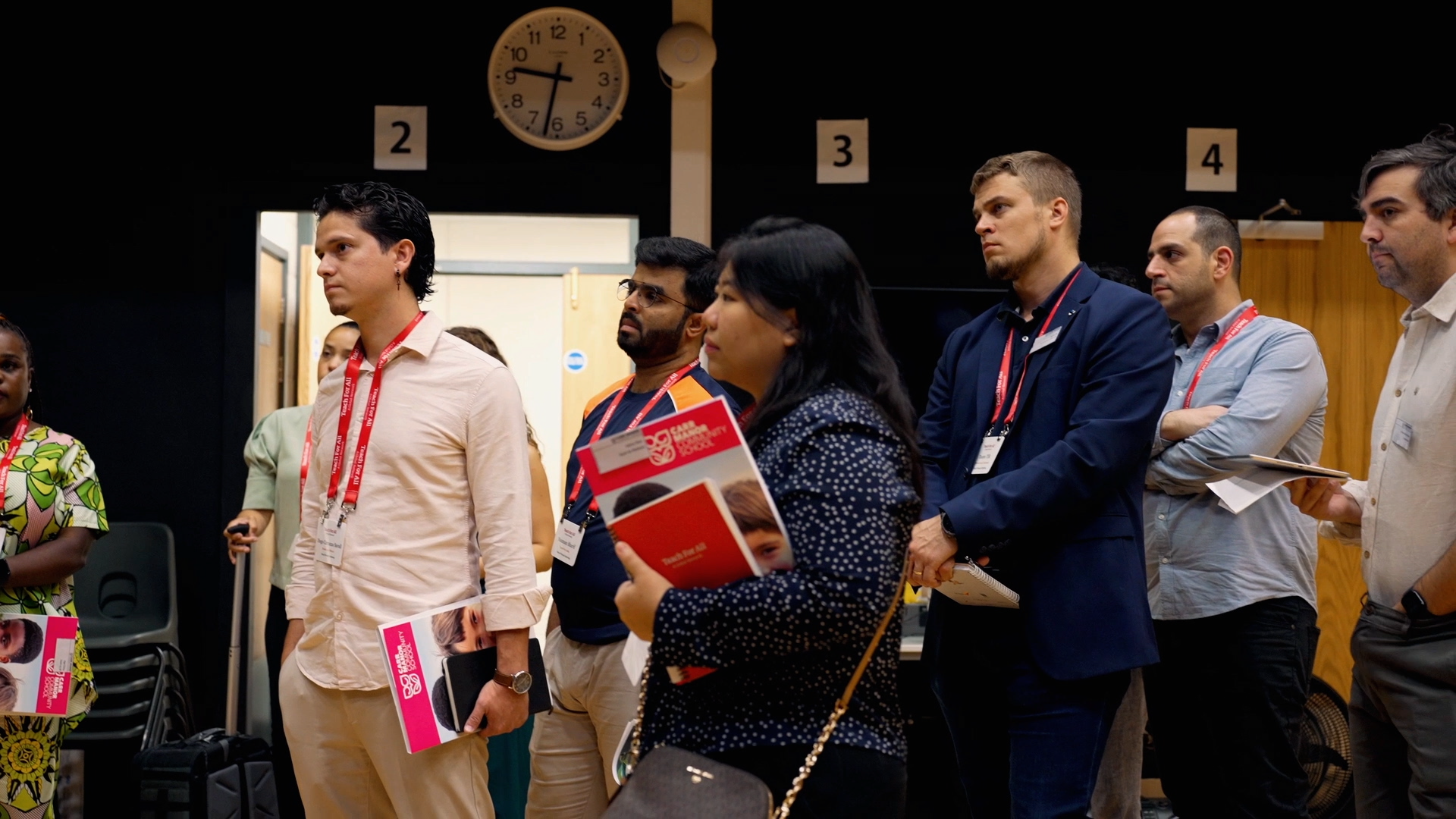 Preview image for the video "The Global Community of School Principals at Teach For All" - A diverse group of adults holding notebooks and wearing nametags stands in a school gym attentively looking out of frame as if someone is speaking to them.
