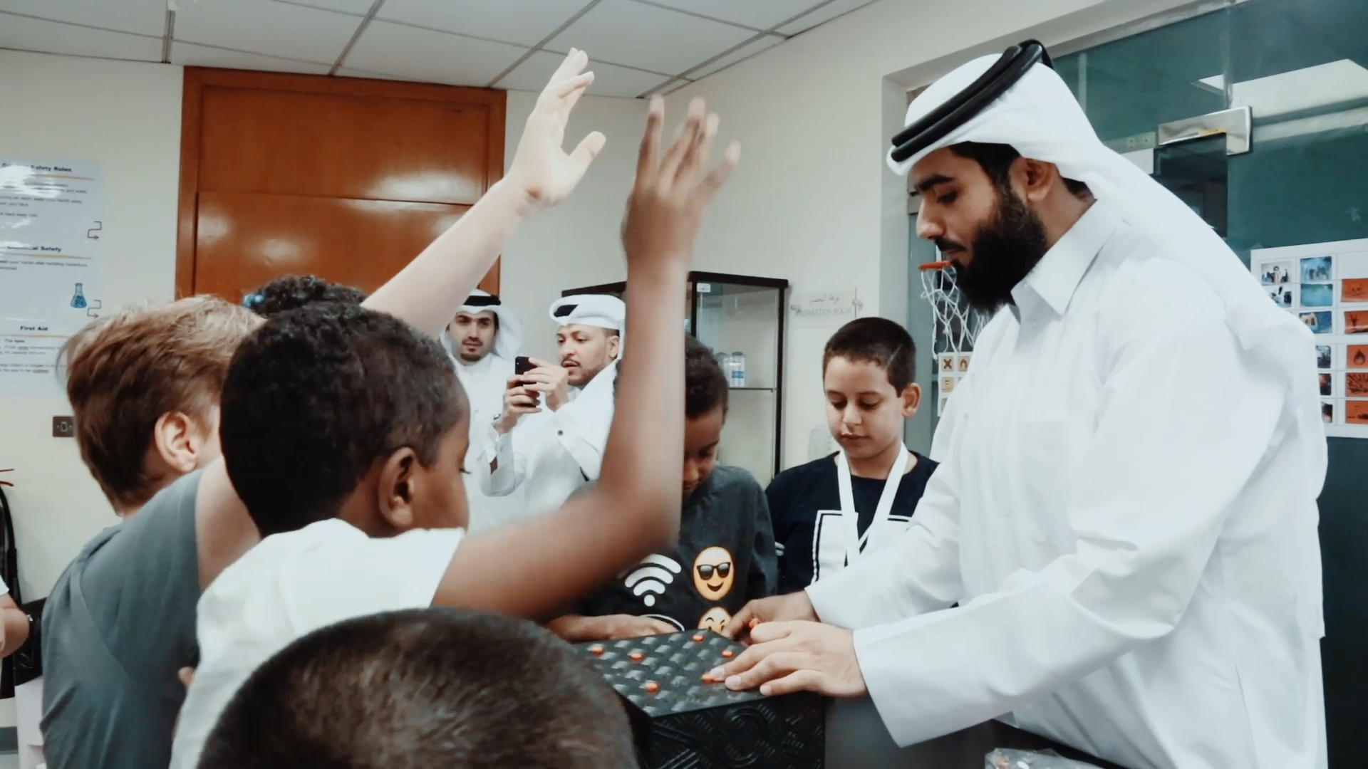 Preview image for the video "Network Changemakers: Mohammed Al Janahi - Teach For Qatar" - A young man in a long white robe (thobe) and white headscarf (ghotrah) stands before several boys raising their hands.