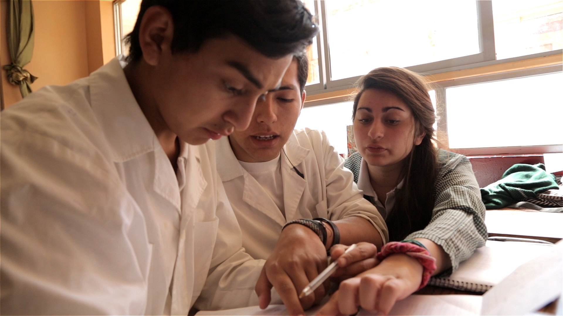 Preview image for the video "Empowering Future Leaders: The Future of Work Initiative at Teach For All" - A young woman with long brown hair and two young men with brown hair and tan skin wearing white lab coats work on a problem at a desk.