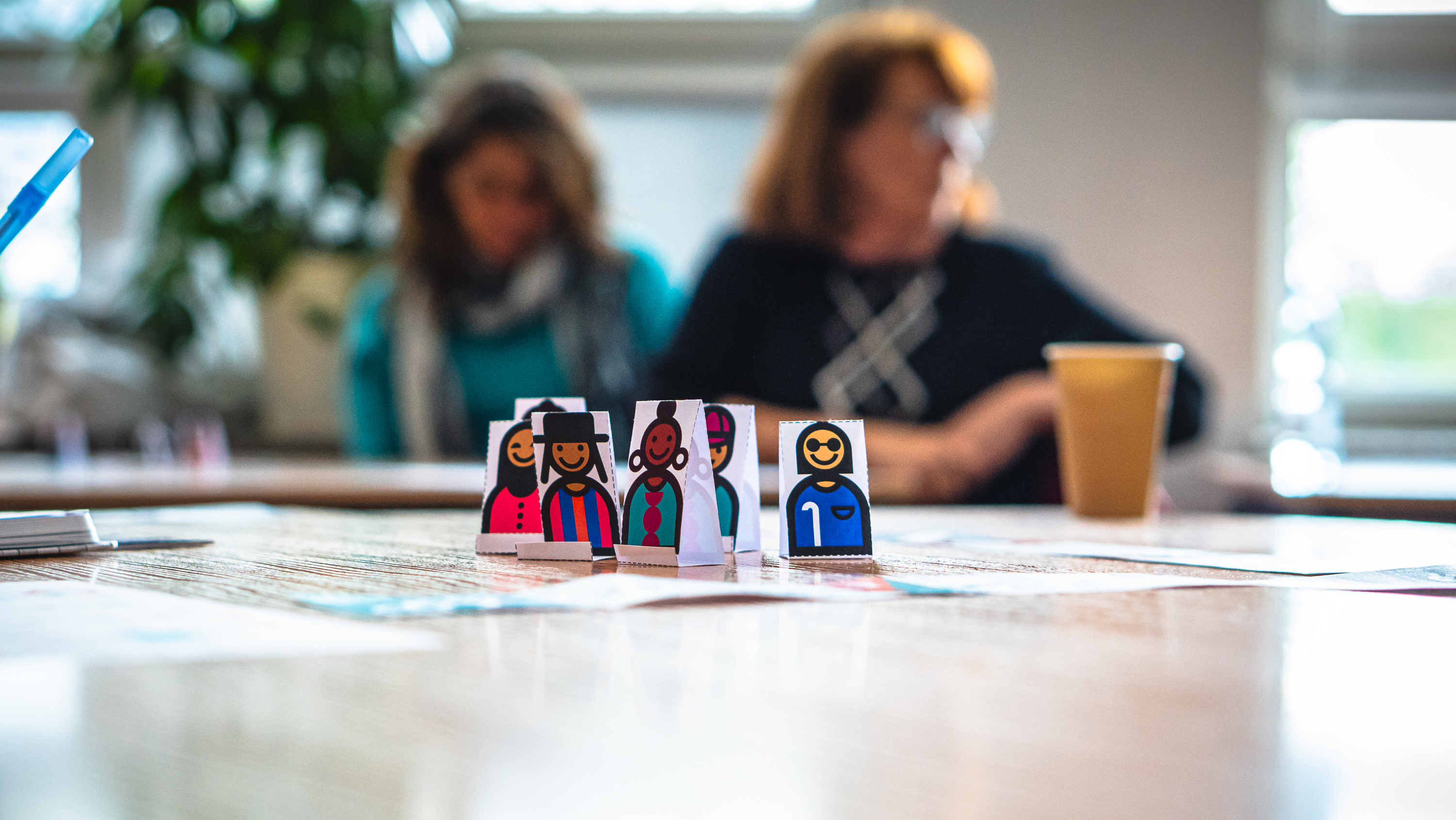 In the foreground are handrawn paper figures of diverse humans on a table while two adult women are visible out of focus in the background seated at another table