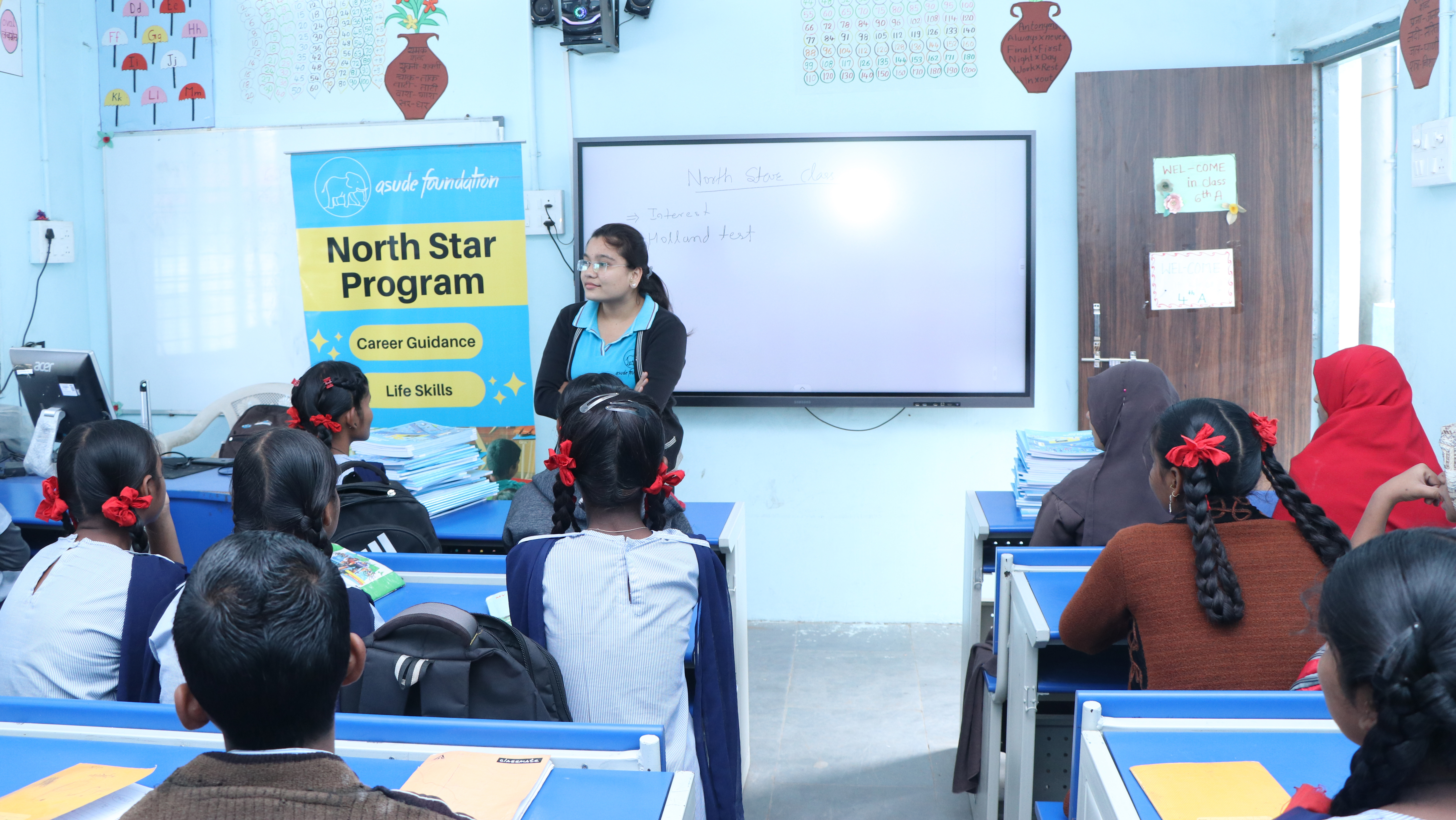 A young woman stands in front of a classroom of school children next to a banner for Asude Foundation's North Star Program