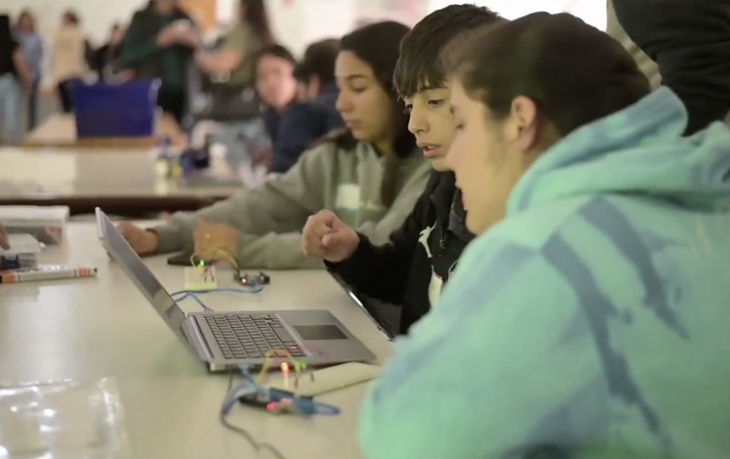 Three teens look at a laptop computer at a table, the girl closest to the camera wears a tie-dyed green sweatshirt.