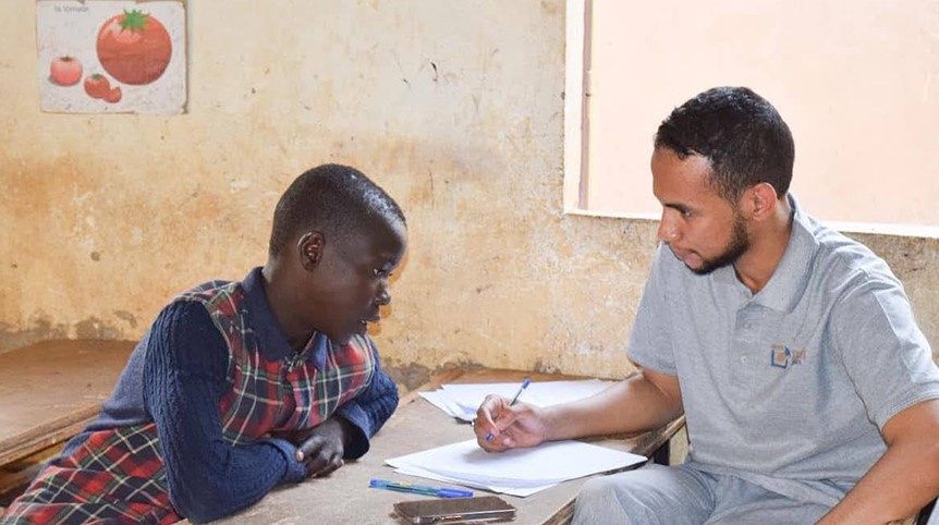 A black girl in a plaid school uniform and a young Black man in a grey Teach For Niger polo shirt look at a notebook together