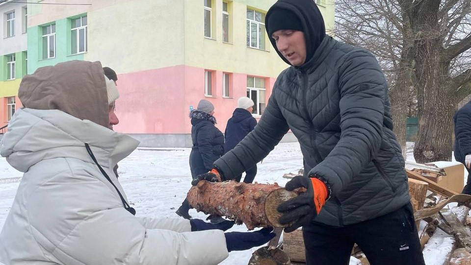 A woman in a black winter jacket and hood hands a log to another person in a light grey winter jacket and hat