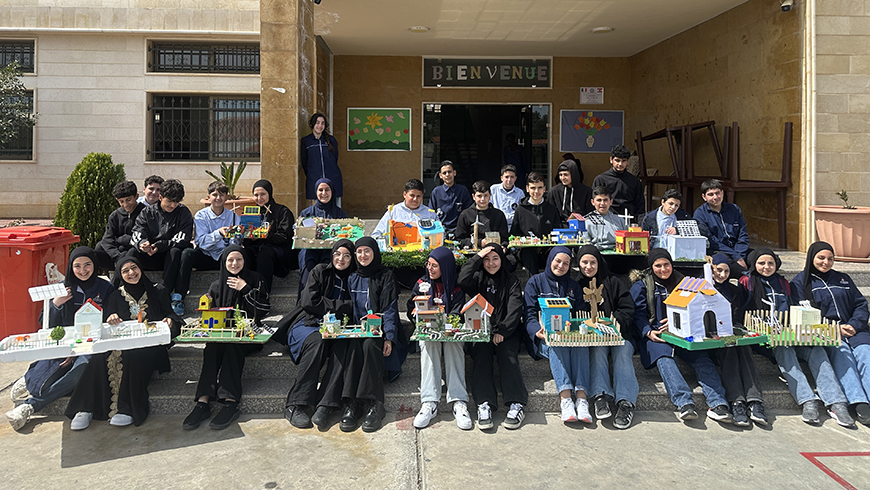 Many children in school uniform sit on steps outside their schools holding models of homes
