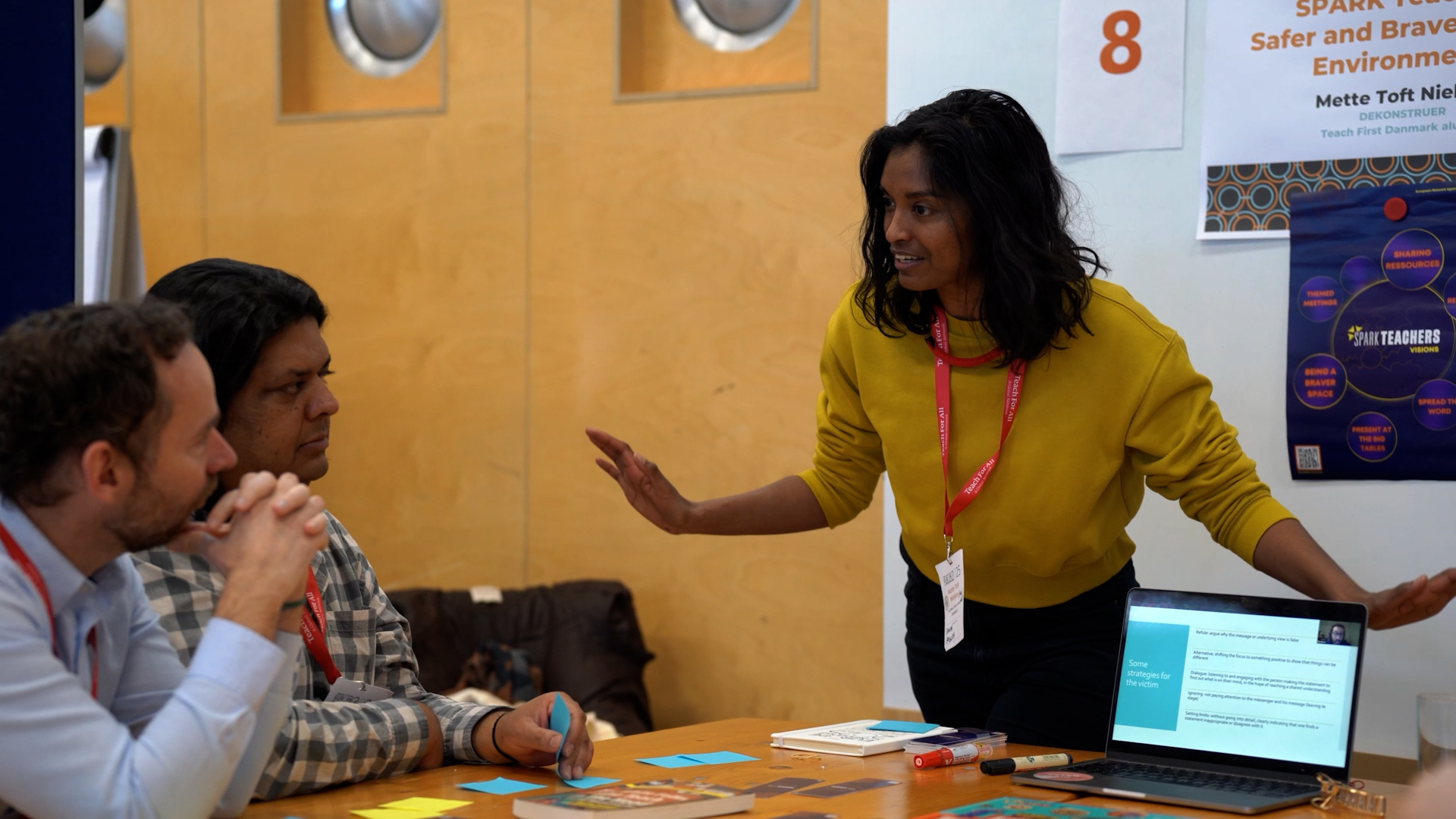 Preview image for the video "In Denmark, Mette Toft Nielsen is Advancing Anti-Racism in Education" - A woman with brown hair and brown skin in a yellow sweatshirt gestures with both hands as she speaks to two men seated at a table during a workshop presentation..