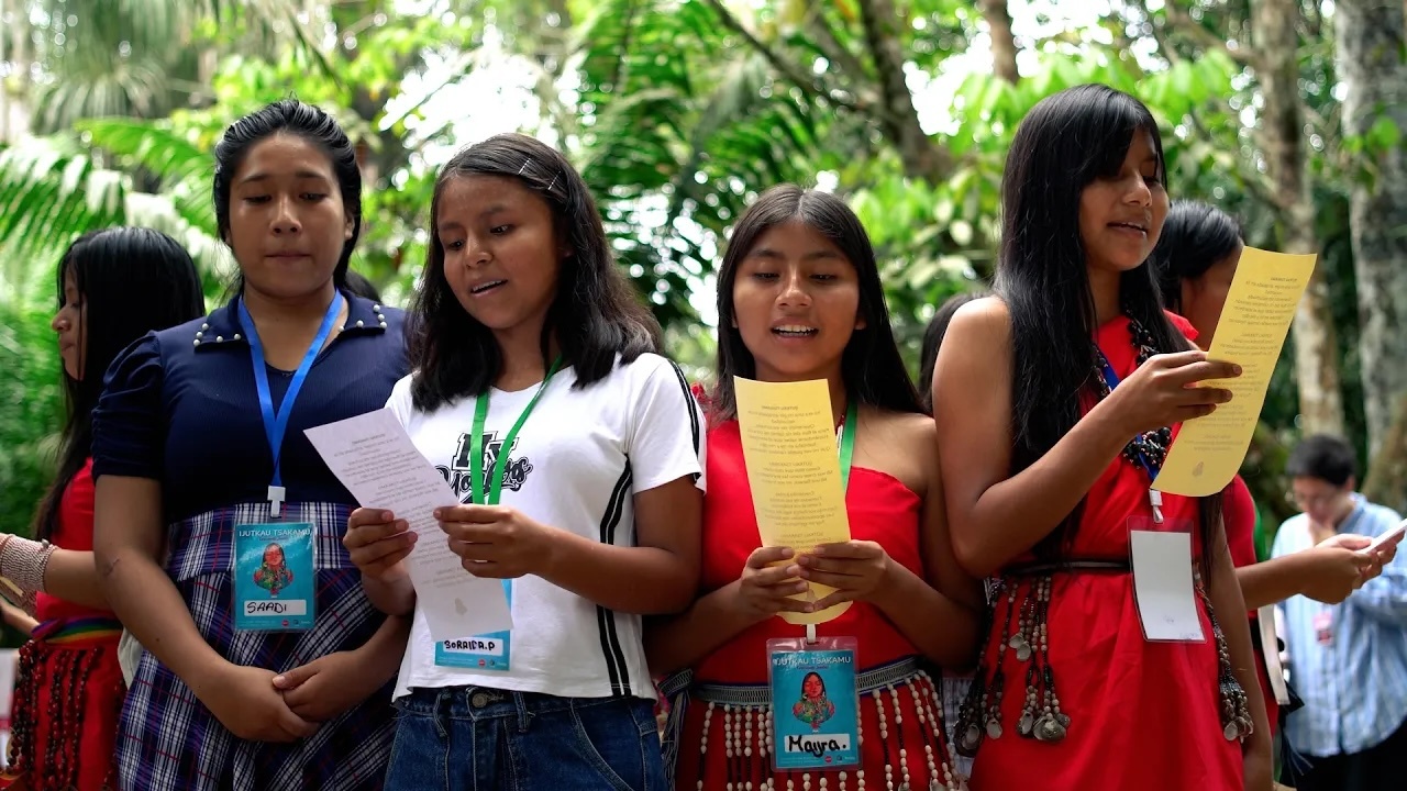 Preview image for the video "Voices of Amazonas: Partnering with communities to redefine leadership by empowering girls" - A group of young Indigenous women stand together outdoors surrounded by lush greenery, holding printed papers as they sing or read aloud during an event..
