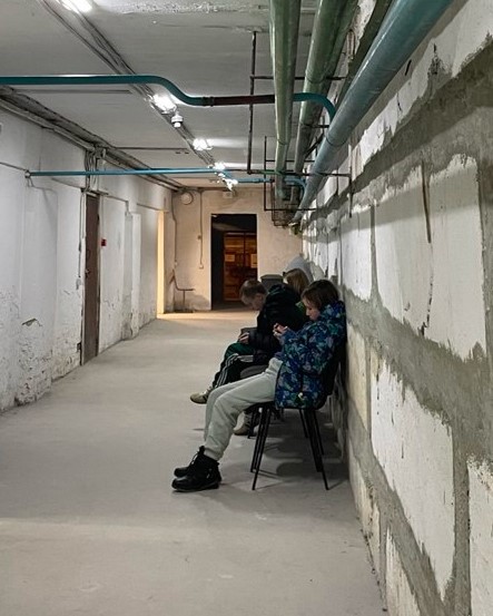 Young teens in winter coats sit against a raw concrete wall in a bomb shelter with pipes on the ceiling