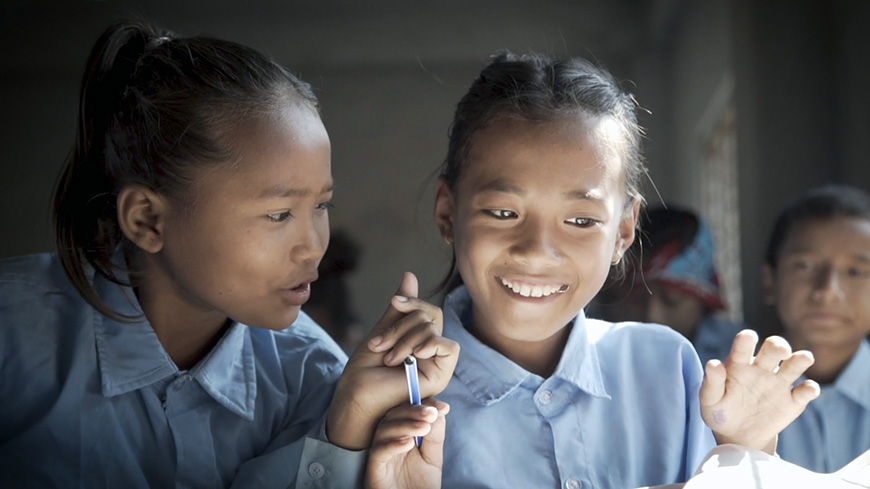 Preview image for the video "Teach For All - A Community of Changemakers" - Two Asian girls wearing blue blouses smile while one holds a pen.
