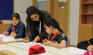 A female teacher wearing a dark headscarf points to a paper in front of a young girl with a braid