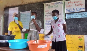 Three women mix materials in plastic basins in front of a chalkboard