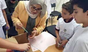 A female teacher wearing a head scarf demonstrates a science activity to two boys and other children who are mostly out of frame