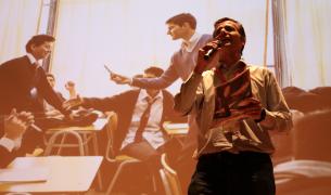 A man speaks into a microphone with an image of a teacher in a classroom behind him