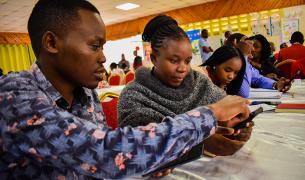 A young Black man points to something on a digital device in a young black woman's hand