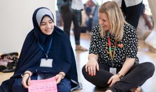 A young student in a hijab sits on the floor next to a blonde, white woman. They are smiling and talking