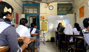 Students seated at desks in a classroom, shot from behind their heads as they look at a female teacher presenting in front of the whiteboard