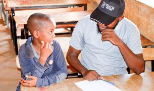 A young Black man in a baseball cap that says Teach For Niger and a young Black boy smile at each other while sharing a wooden desk in a classroom