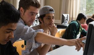 A young man points at a computer screen with teenage boys on either side of him, one in baseball cap