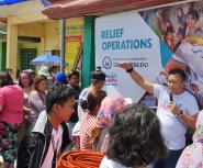 A group of people listen to man holding a microphone speaking in front of a large banner saying "RELIEF OPERATIONS"