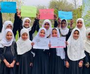 A group of young girls wearing white hijabs stand outdoors, holding up colorful signs that read "Happy 10th Anniversary Of Teach For Afghanistan."