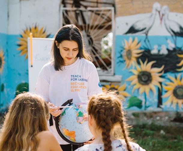 A female teacher with long brown hair shows a globe to two female students we see from behind, with a backdrop of outdoor walls painted with sunflowers