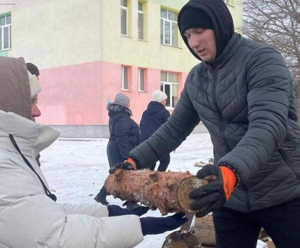 A woman in a black winter jacket and hood hands a log to another person in a light grey winter jacket and hat