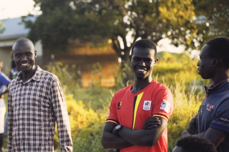 Three African men smile as they stand together in a grassy area with a big tree in the background