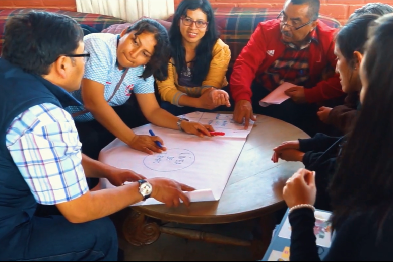 A group of adults of various ages surround a table collaborating on a project