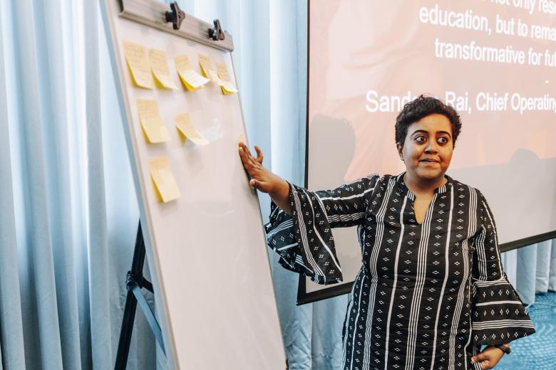 An woman with brown skin and short hair wearing a black and white patterned dress points to a whiteboard covered with several post-its