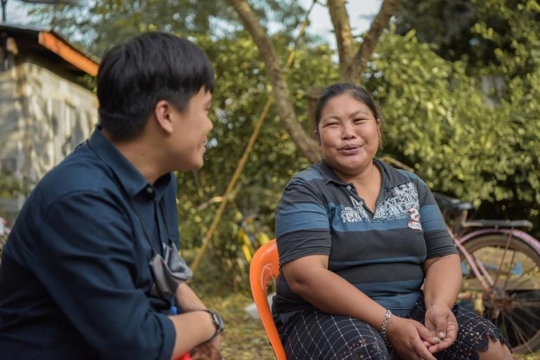 An Asian woman sits outside in an orange chair smiling at a young Asian man sitting next to her. He's facing her so we only see the side of his head