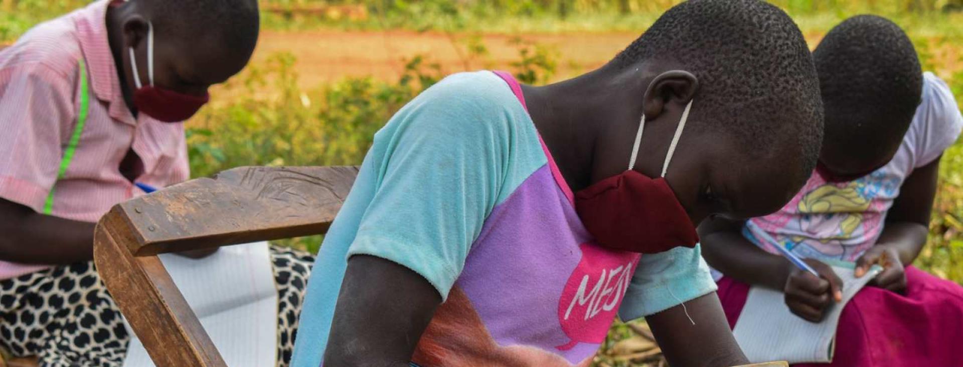 Three children writing in notebooks, outdoors, wearing facemasks