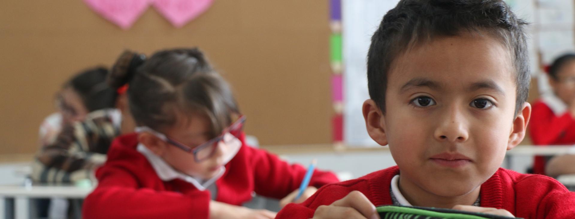 A boy in a red sweater with an open notebook faces the camera while a girl behind him also in a red sweater writes at her deesk. On the chalkboard behind them are two pink hearts.