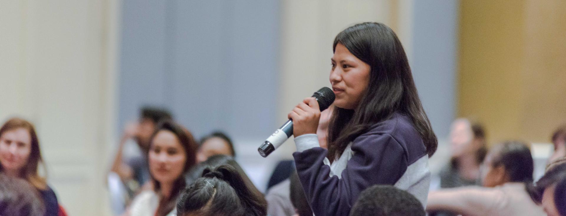 A young South Asian woman stands in an audience and speaks into a microphone