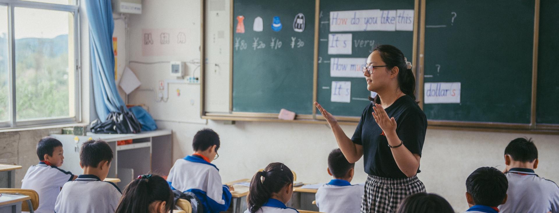 A young Asian woman stands in a classroom in with a chalkboard behind her among students wearing white shirts seated in desks 