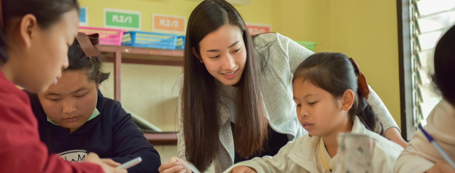 A young Asian woman with long hair leans over a table where three Asian girls are writing in workbooks