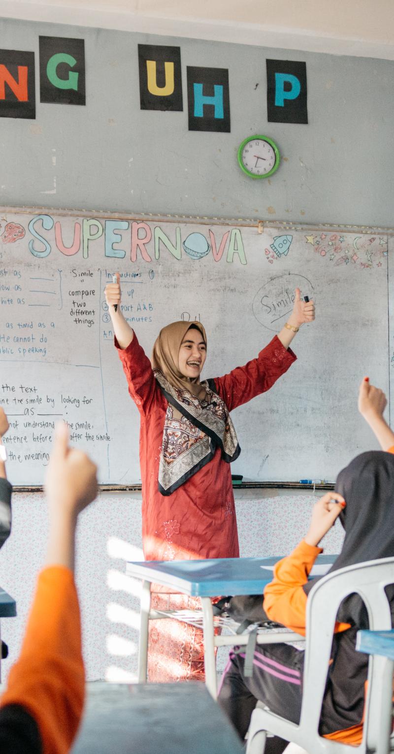 A female teacher in a red dress and head scarf stands in front of a white board facing her students in desks. Everyone raises their arms giving a thumbs-up