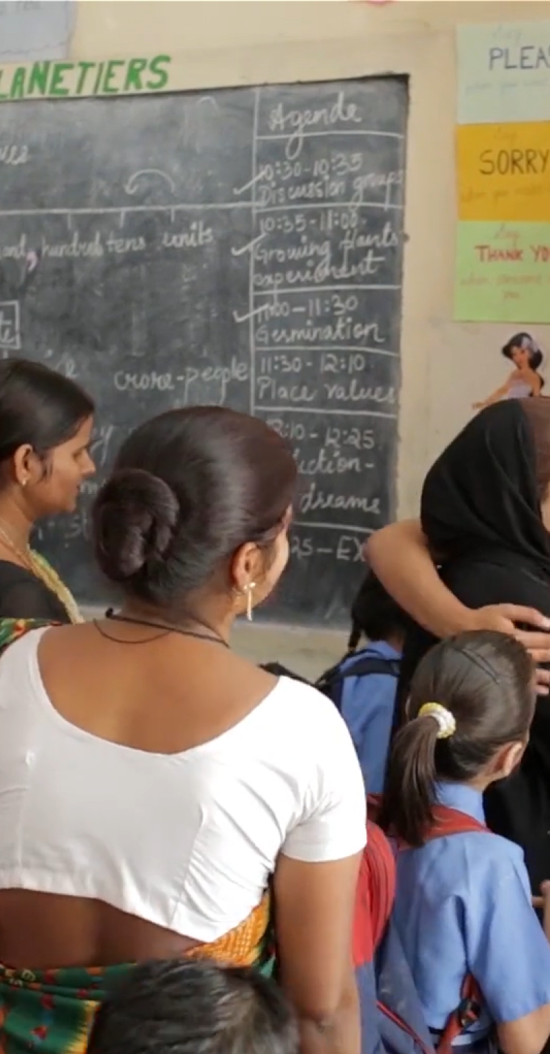 In a classroom, a young Indian woman in a red dress has her arm around another woman with a head scarf, surrounded by other women and girls in school uniform