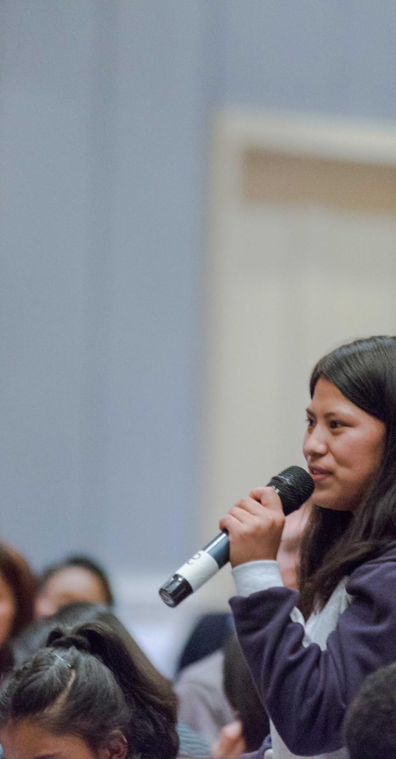 A young South Asian woman stands in an audience and speaks into a microphone