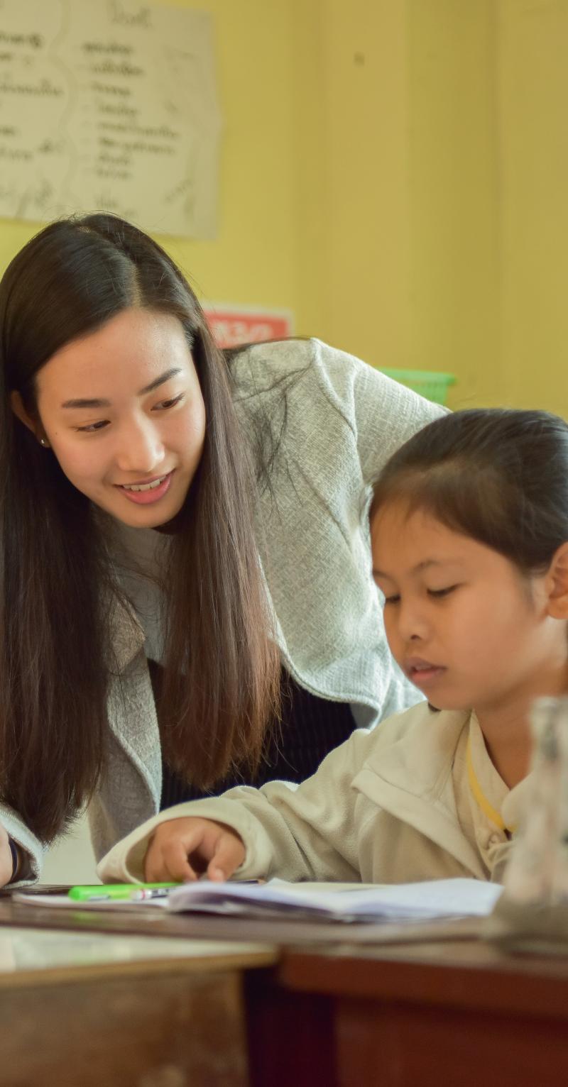 A young Asian woman with long hair leans over a table where three Asian girls are writing in workbooks