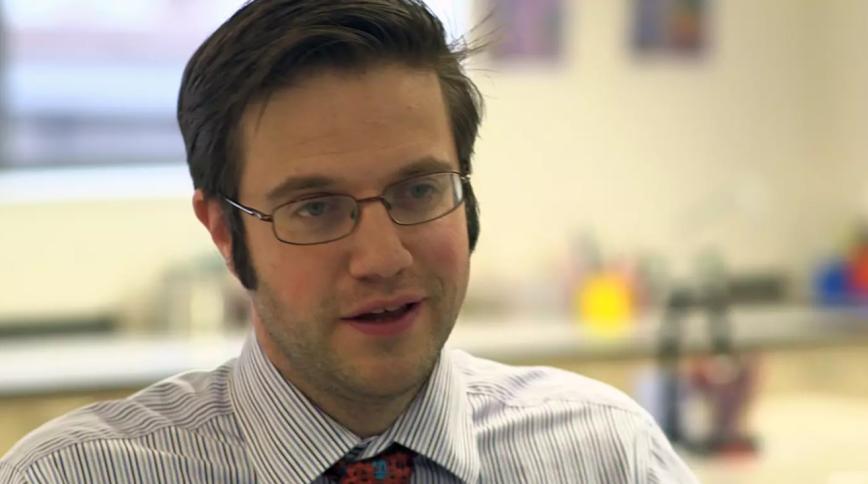 Close up headshot of a young white man with brown hair and glasses wearing a shirt and tie