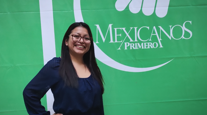 A young woman with long brown hair and glasses stands in front of a green sign that says Mexicanos Primero