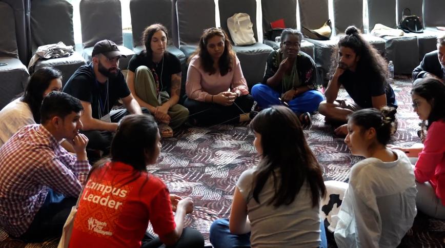 A diverse group of young adults sit in a circle on a carpeted floor