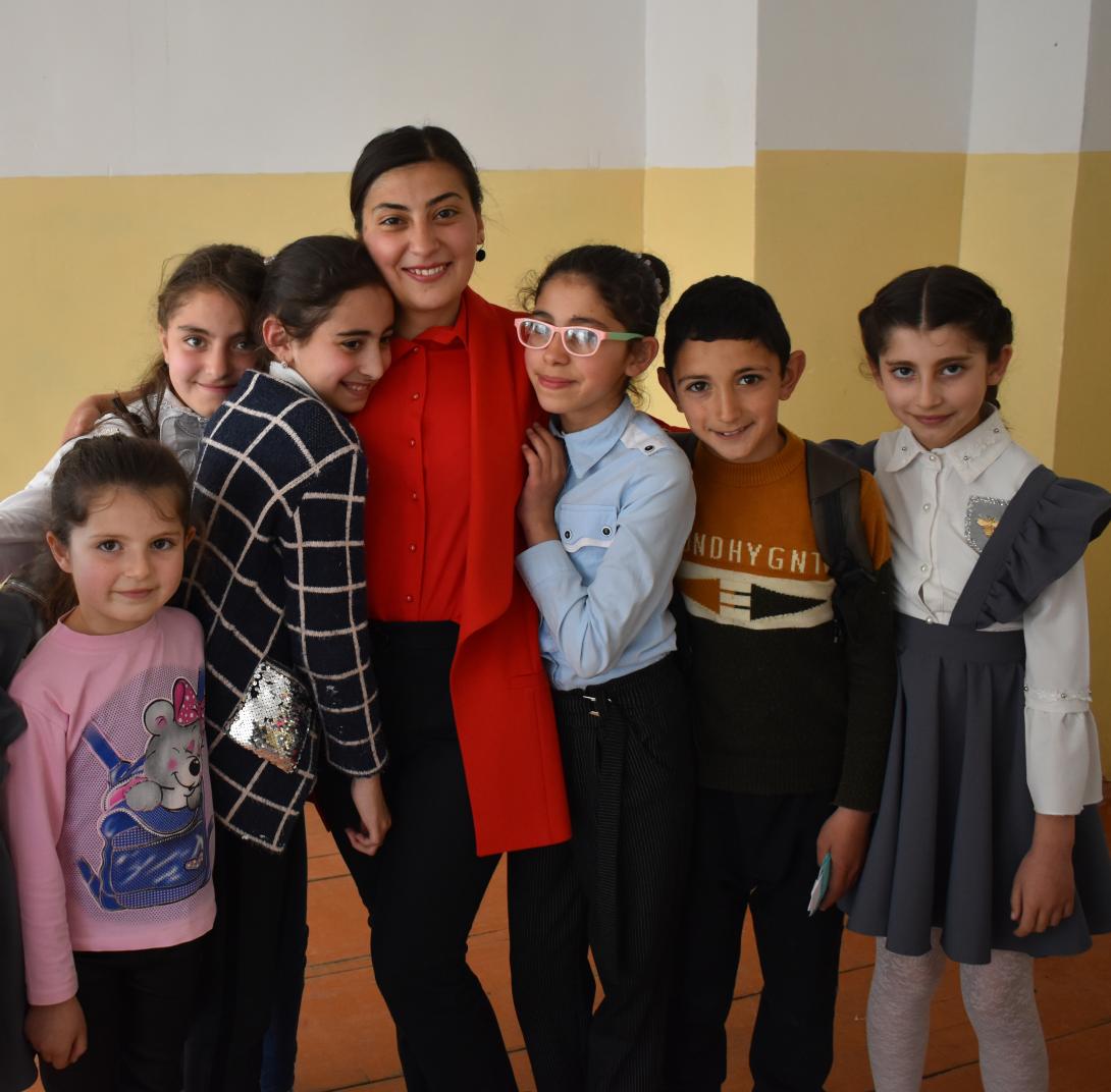 A female teacher in a red shirt and jacket has her arms around six students, one of whom rests her head on the teacher's shoulder