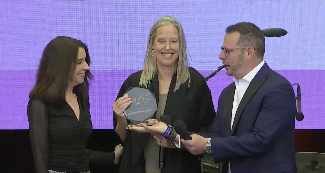 A white woman with long blond hair receives an award from a white man in a suit and a white woman with brown hair 