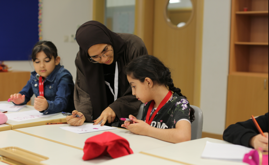 A female teacher wearing a dark headscarf points to a paper in front of a young girl with a braid