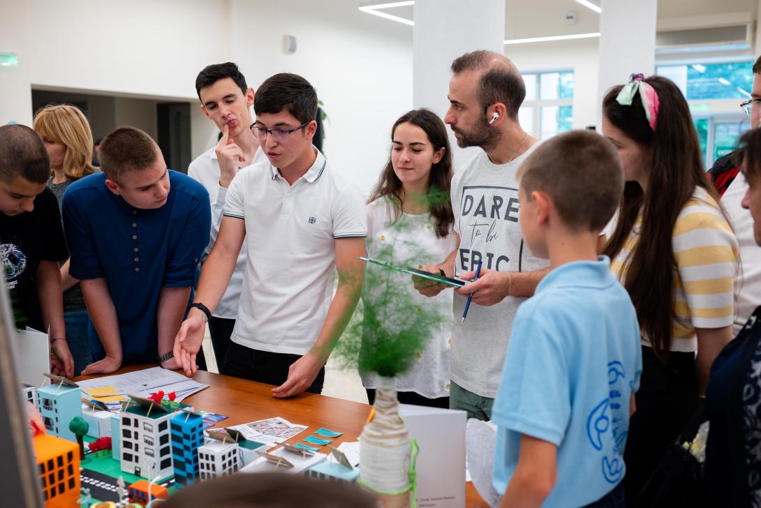 A teenage boy points to a colorful model of a city on a table while surrounded by several other teenagers and a man with a clipboard, all of whom are listening attentively